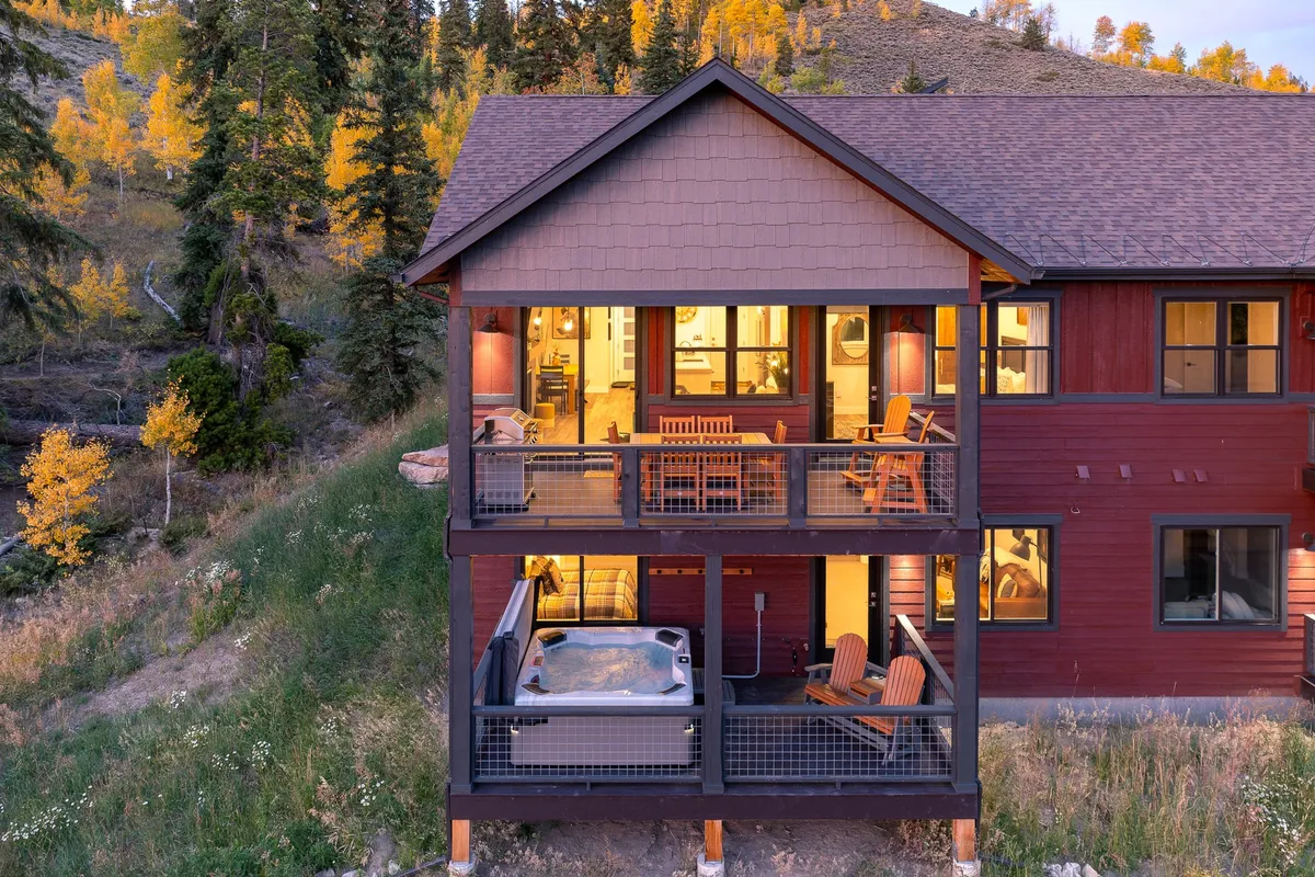 This scene features a covered patio with a bubbling hot tub, deck furniture, and large windows looking out to surrounding trees. The cozy arrangement and soft lighting create an inviting atmosphere perfect for relaxation or après-adventure gatherings.