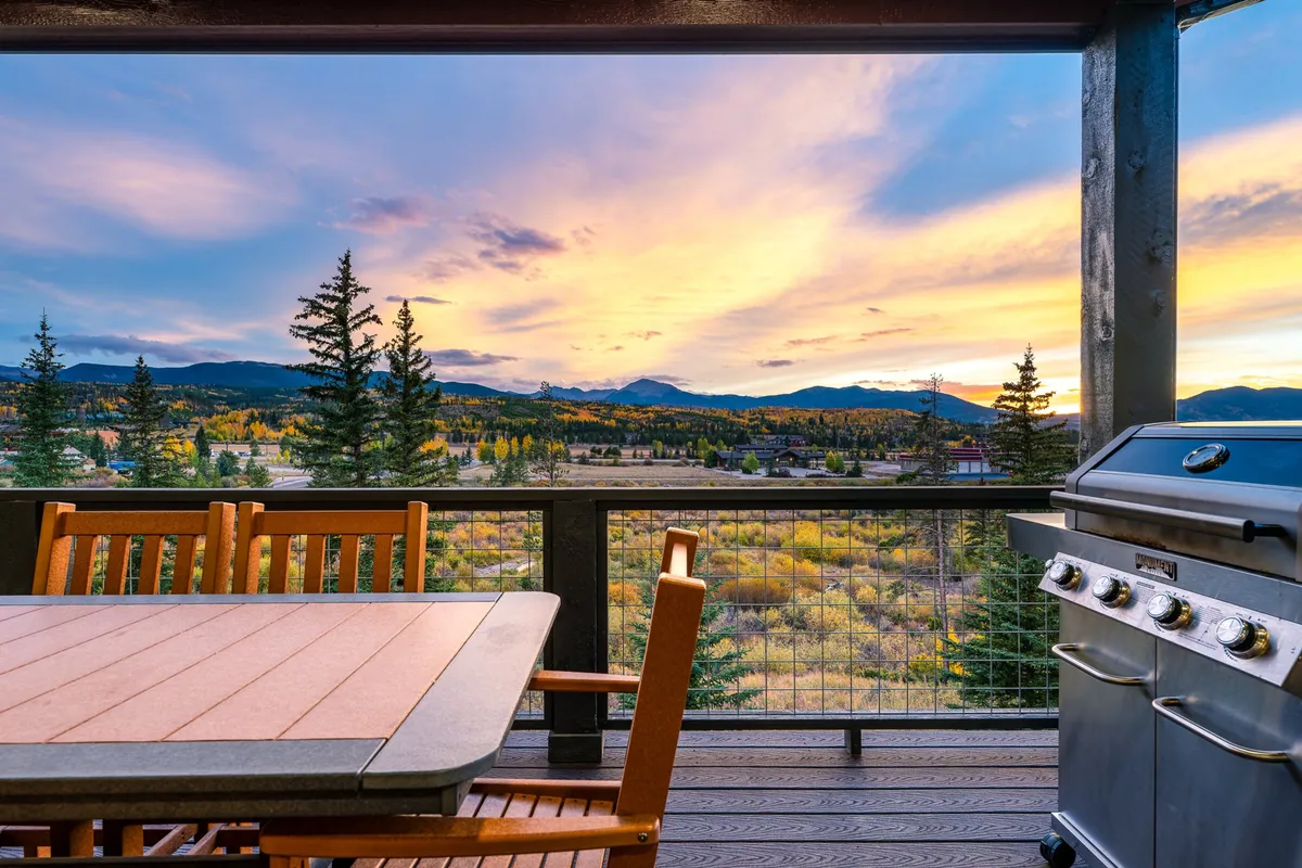 An inviting outdoor dining area features a table set for a meal, a grill ready for barbecuing, and breathtaking mountain views at sunset. Trees frame the space, creating a tranquil atmosphere for socializing and unwinding after a day of productivity.