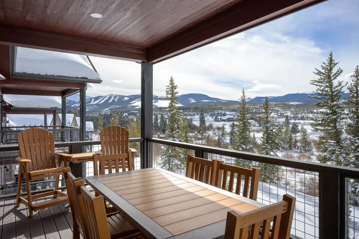 A spacious covered deck furnished with a wooden dining table and Adirondack chairs overlooks a snowy Rocky Mountain landscape, with Byers Peak visible in the distance. The scene captures a serene outdoor dining area perfect for morning coffee or relaxing after a day of adventure.