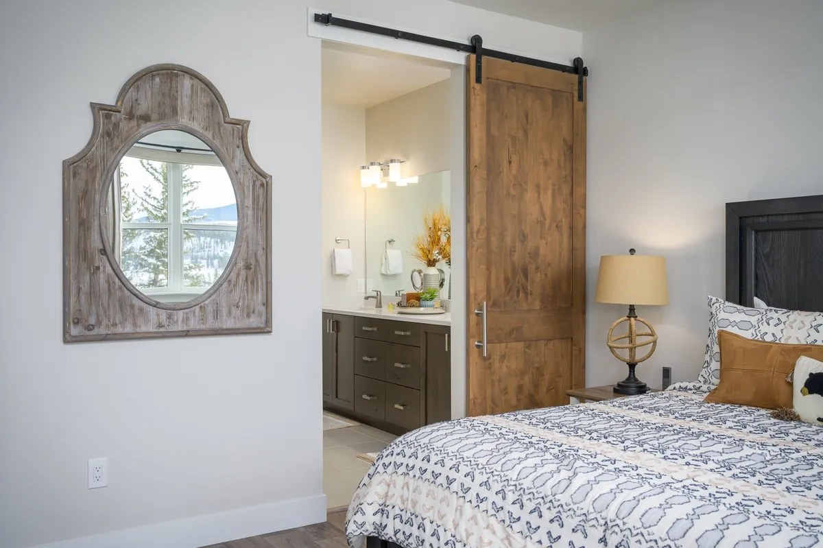 A cozy king bedroom with patterned bedding is shown, featuring a rustic wooden barn door that leads to an ensuite bathroom. The image also includes a decorative framed mirror and a glimpse of the bathroom vanity area, blending the bedroom and bathroom spaces.