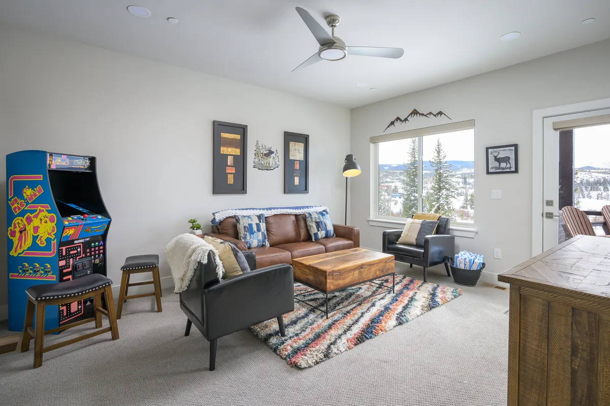 A vibrant lower-level lounge with a leather sofa positioned near a coffee table and a classic arcade game. The large window offers natural light and a scenic view, making the space both playful and relaxing. The setup invites group gatherings, game nights, and moments of relaxation, matching the fun, multi-generational spirit described in the section.