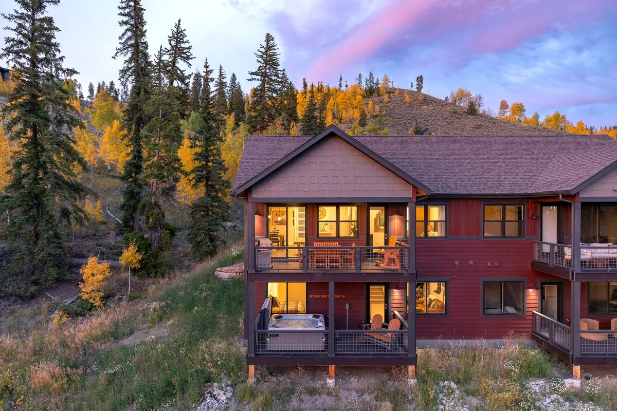 The image shows the exterior of a luxurious chalet in Fraser, Colorado, featuring a spacious wooden deck with comfortable seating, a bubbling hot tub, and large windows glowing with warm interior lighting. The deck overlooks scenic mountain views, with the sunset casting a golden glow on the landscape, evoking a peaceful and cozy atmosphere. Seasonal foliage adds to the property's inviting charm.