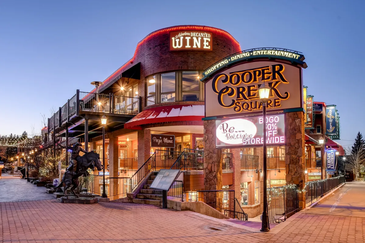 The vibrant Cooper Creek Square sign sets the scene for a lively downtown district, flanked by decorative wine signage and outdoor seating areas. The scene buzzes with the promise of dining, shopping, and entertainment in a communal, year-round environment.