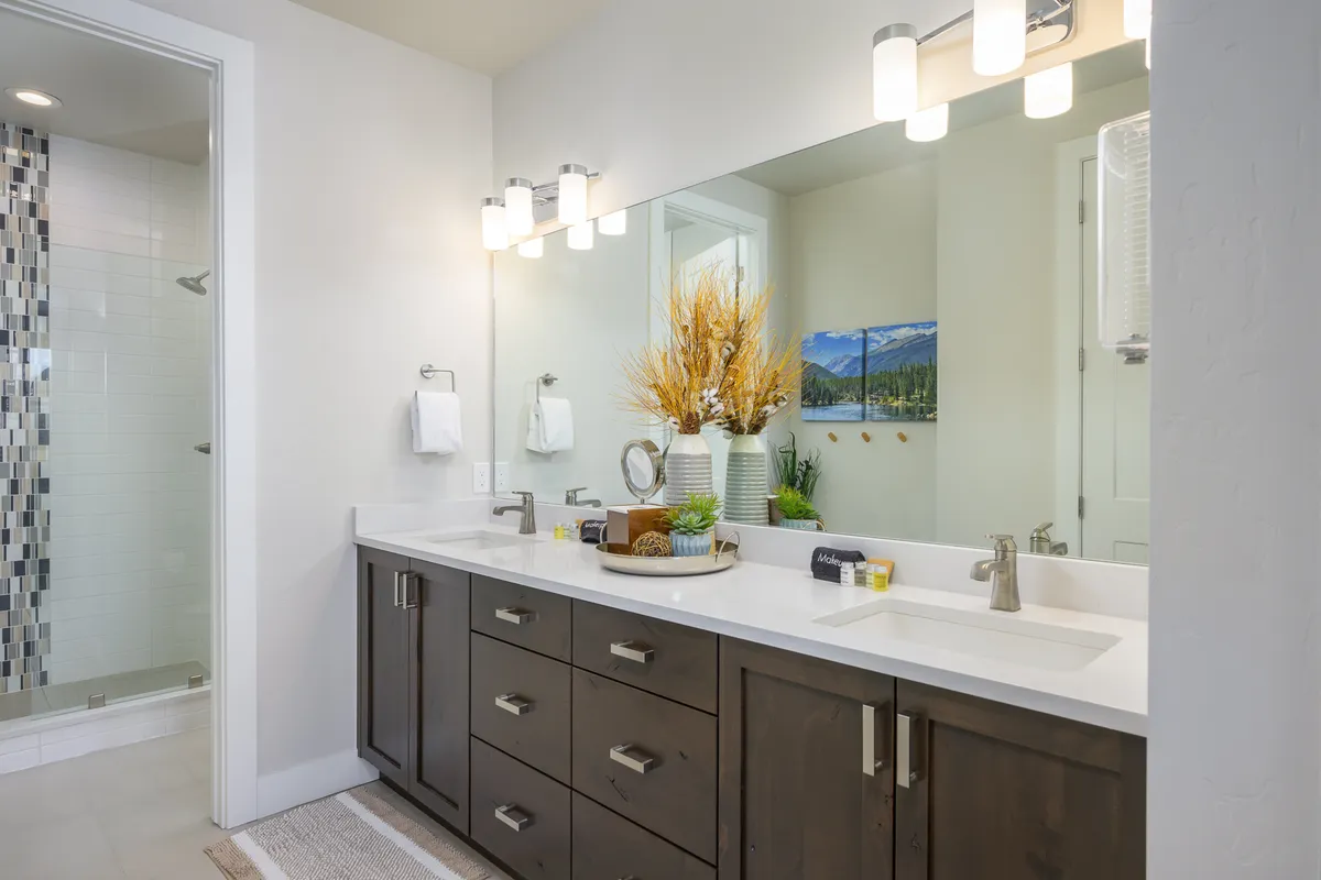 A modern bathroom is depicted with a double sink vanity, large mirror reflecting soft light, decorative plants, a spacious shower area, and sophisticated wall art. The mood is calm and refreshing, suggesting a spa-like experience.