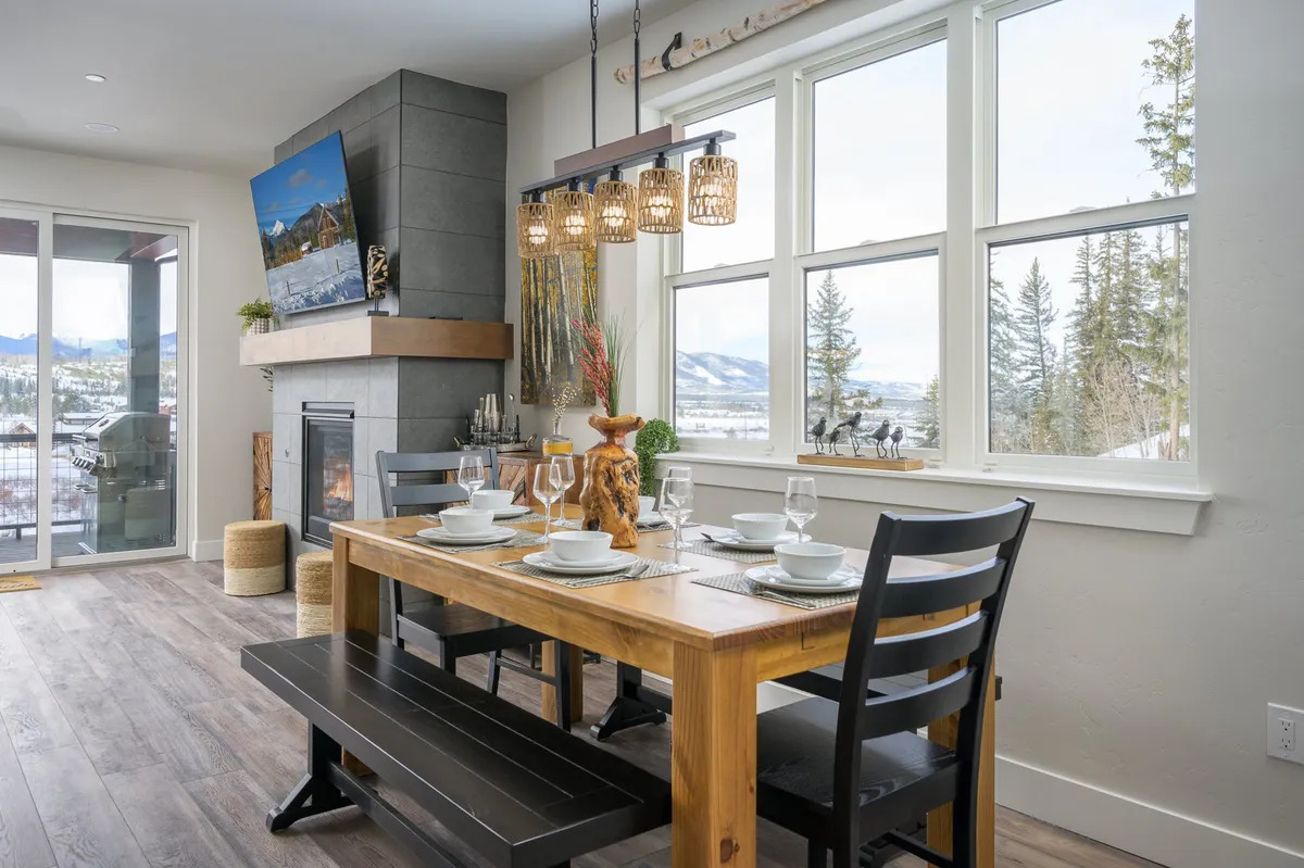 A warmly lit dining area with a central wooden dining table, surrounded by textured chairs. Large windows invite ample natural light and offer sweeping mountain views, while a nearby fireplace adds a cozy ambiance. An elegant chandelier hangs above the table, enhancing the inviting atmosphere.