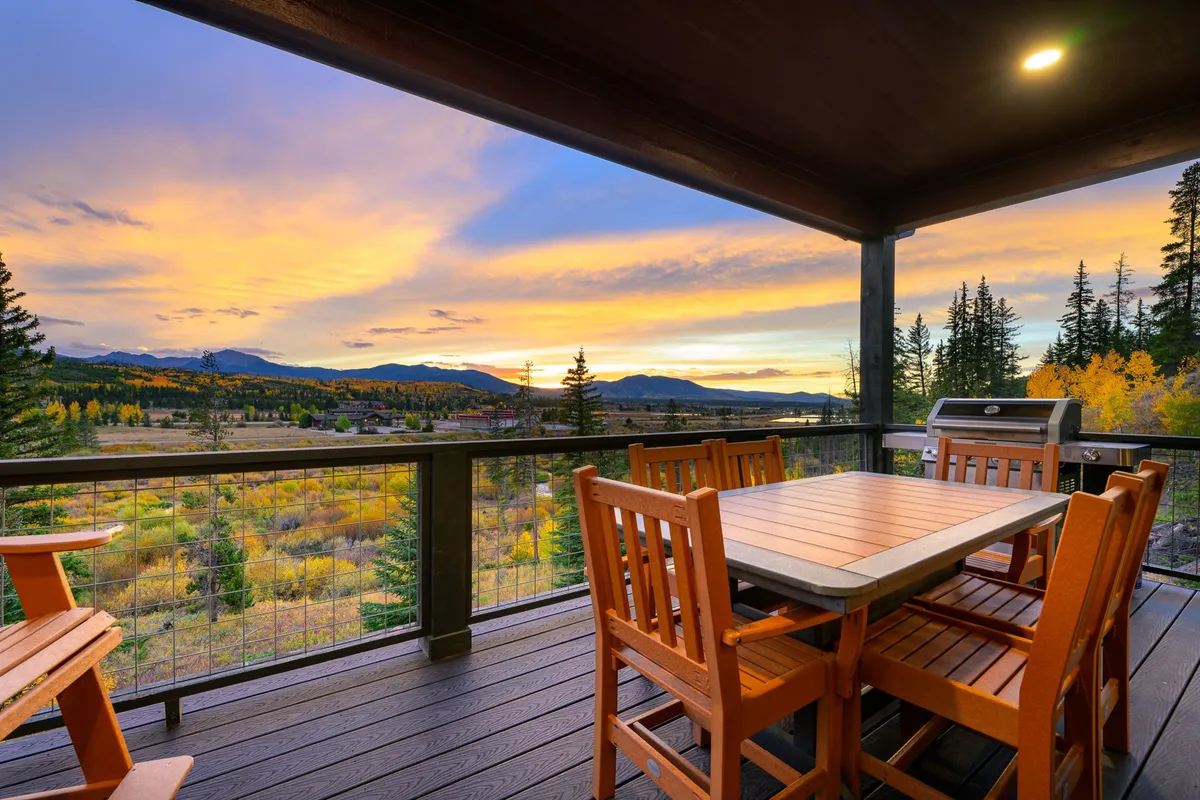 This image captures a wooden outdoor dining set and grill set against a panoramic sunset view with surrounding trees. The scene evokes a sense of relaxation and connection with nature, highlighting the spaciousness and inviting atmosphere of the upper covered deck for alfresco dining and grilling.